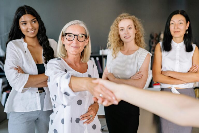 Home Diverse group of women in an office setting shaking hands and smiling, symbolizing partnership.